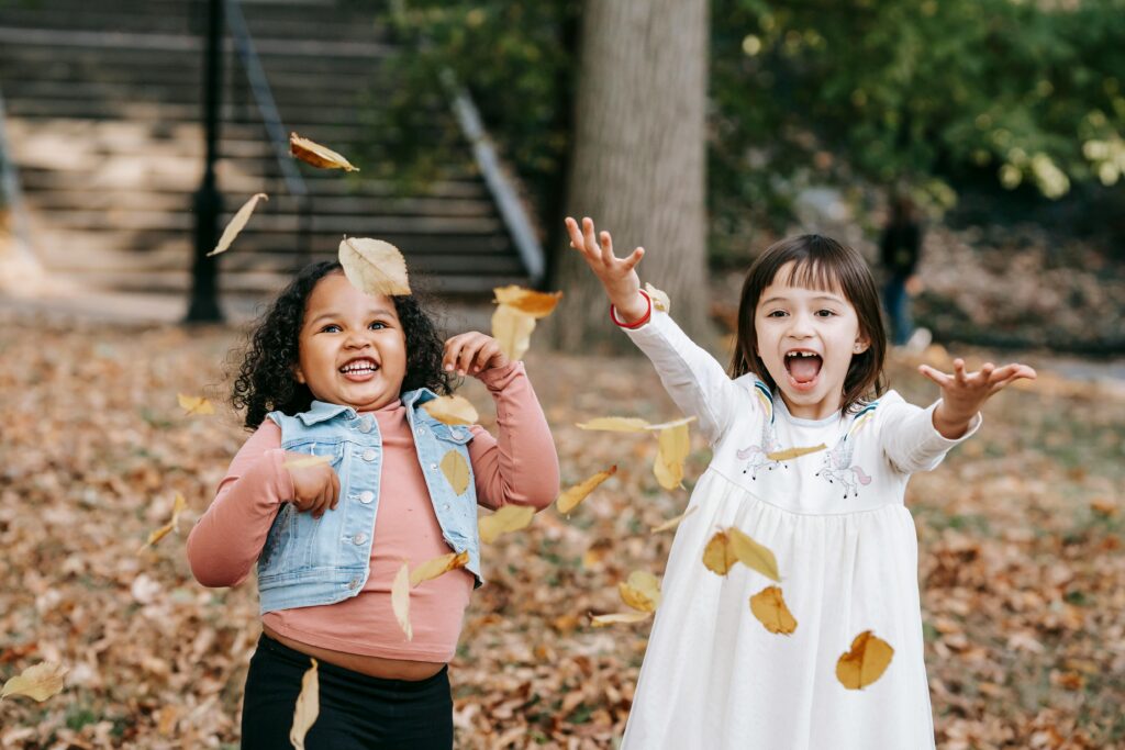 Two cheerful girls toss fall leaves joyfully in the park, capturing autumn fun.