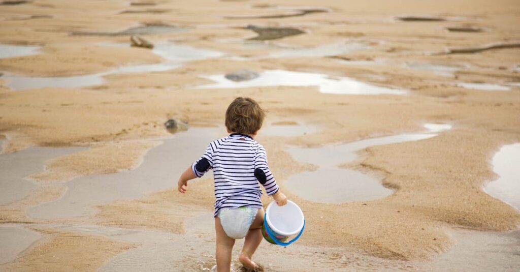 A child carrying a bucket walks along the sandy beach on a sunny day, enjoying leisure time.