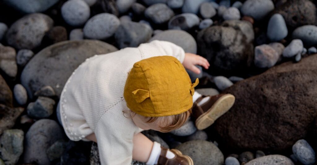 An adorable toddler with a yellow hat sits among large, smooth rocks outdoors.