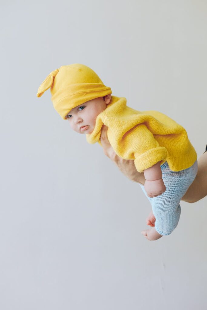 Charming baby in yellow knit outfit held in a playful flying pose indoors.