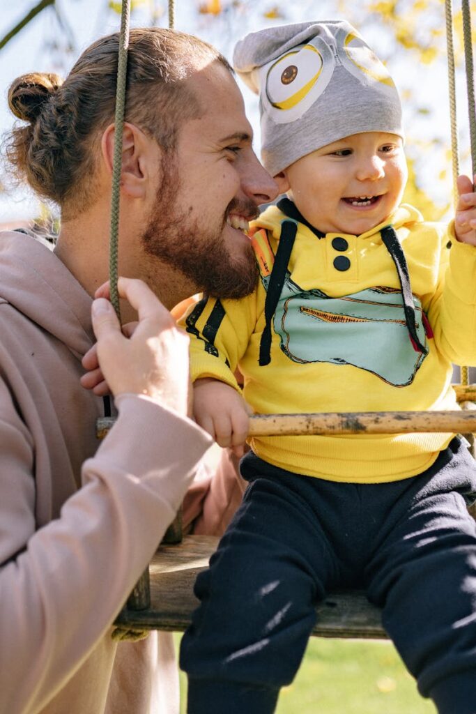 A father joyfully interacts with his toddler on a swing during a sunny fall day outdoors.