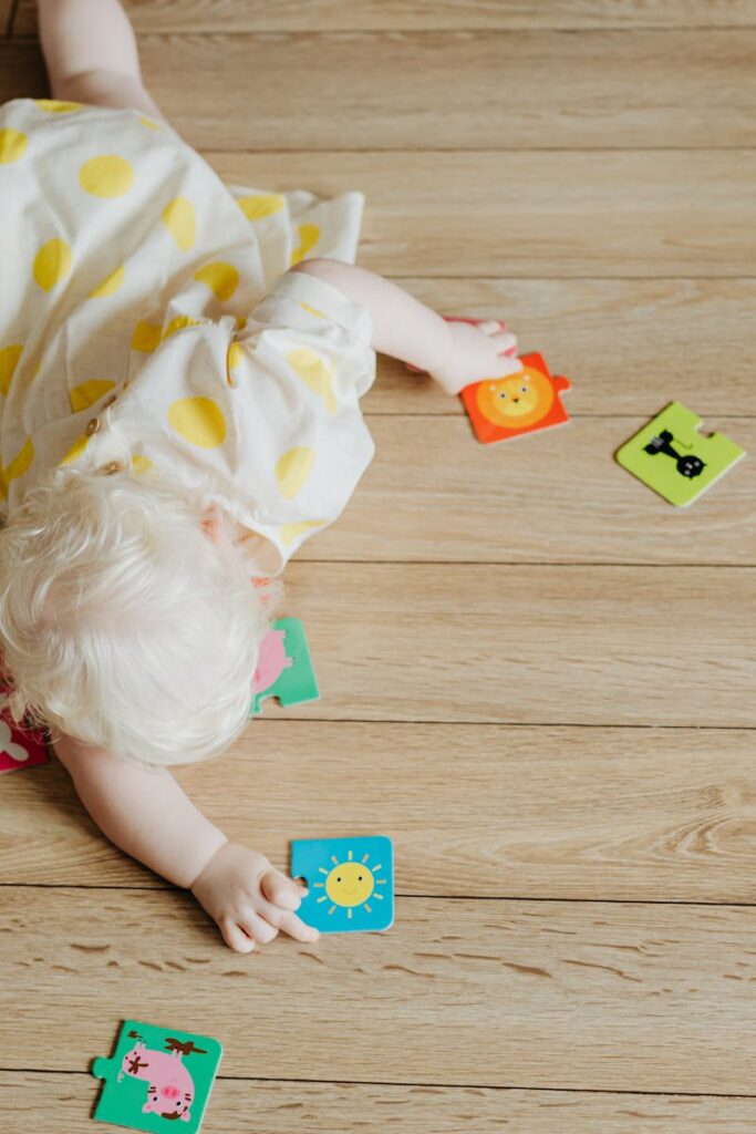 Cute baby in a polka dot dress crawls on wooden floor with colorful puzzle pieces.