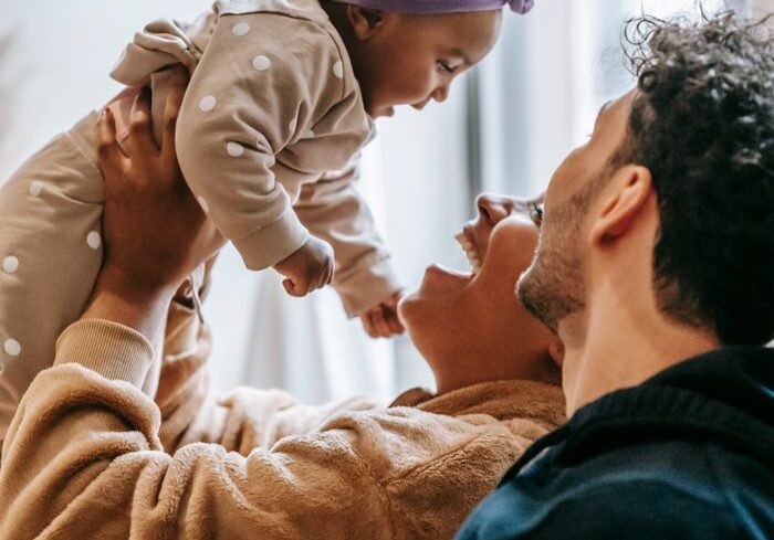 Side view of cheerful ethnic mother lifting cute baby girl while sitting near positive father at home
