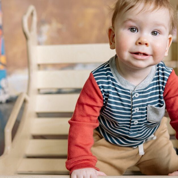Cute toddler with blond hair plays indoors on a wooden toy, wearing colorful clothing, smiling at the camera.